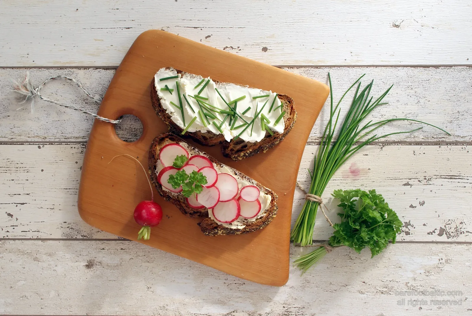 Zwei Scheiben dunkles Roggenbrot mit Biezpiens-Quark, frischem Schnittlauch und Radieschenscheiben auf einem Holzbrett