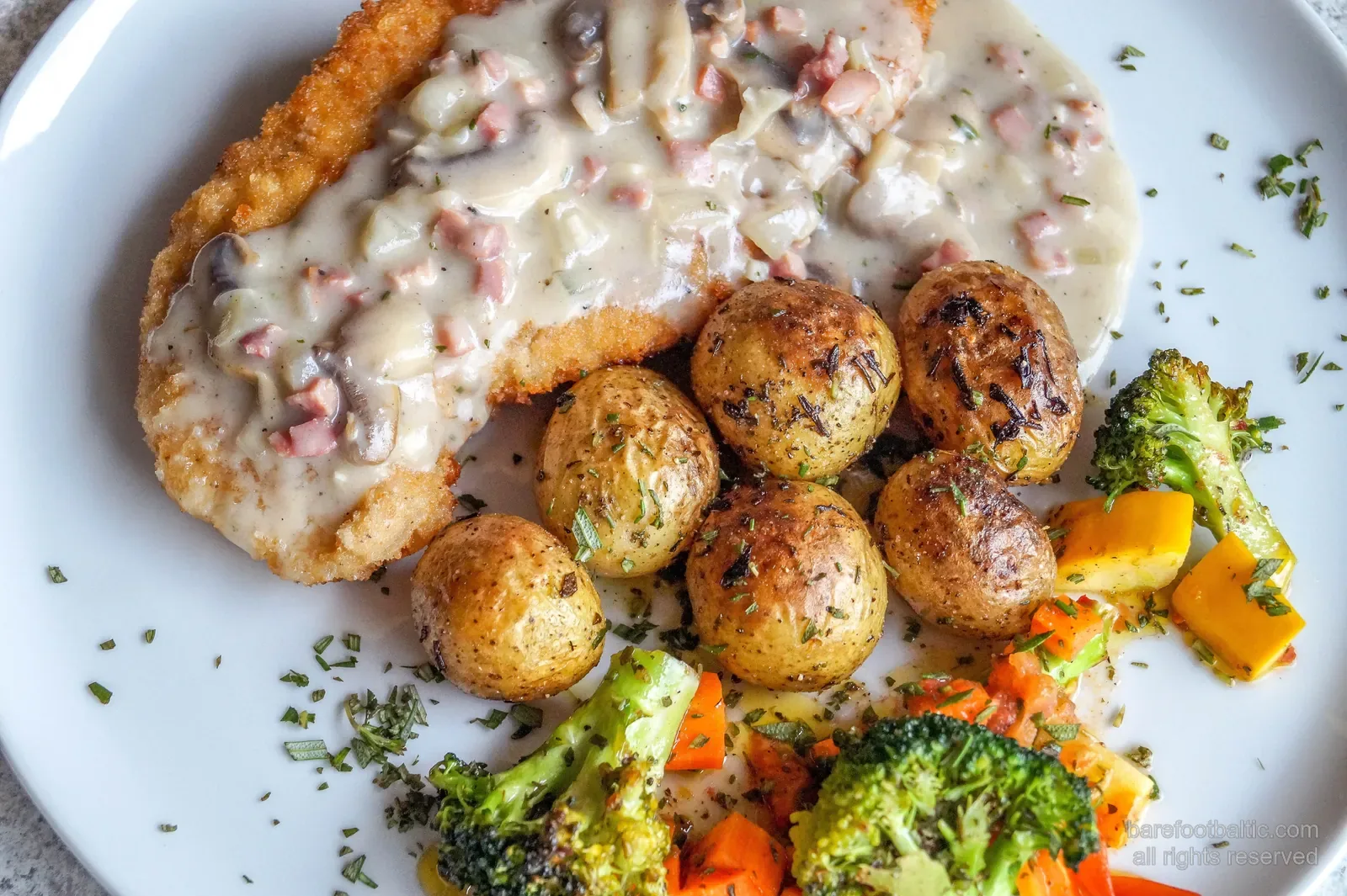 Ein Teller mit Karbonāde &mdash; dem lettischen panierten Schweineschnitzel &mdash; mit cremiger Pilzsauce, Kräuterkartoffeln und Röstgemüse
