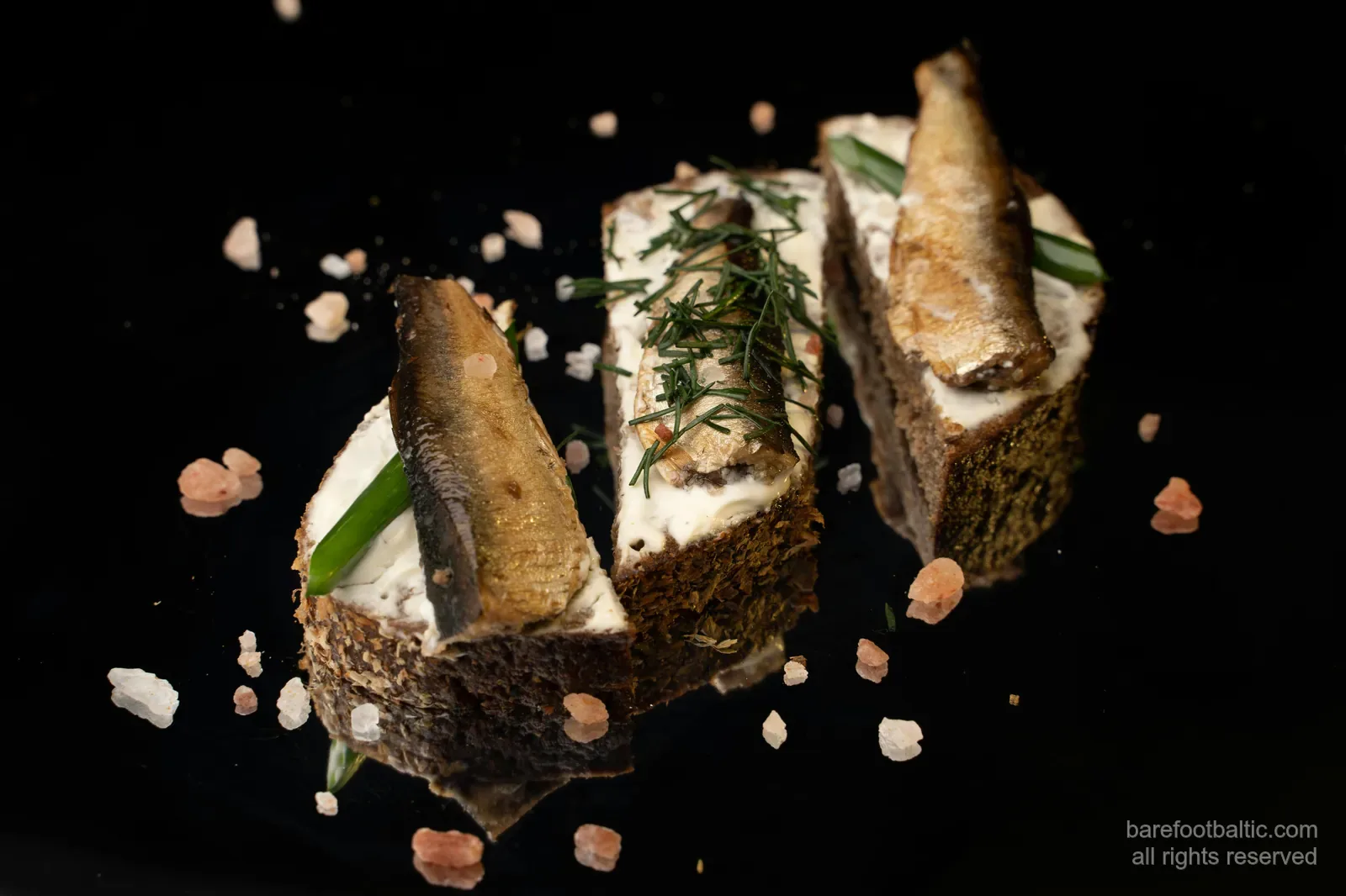 Drei geräucherte Sprotten auf Roggenbrot mit saurer Sahne und Dill, auf schwarzem Hintergrund